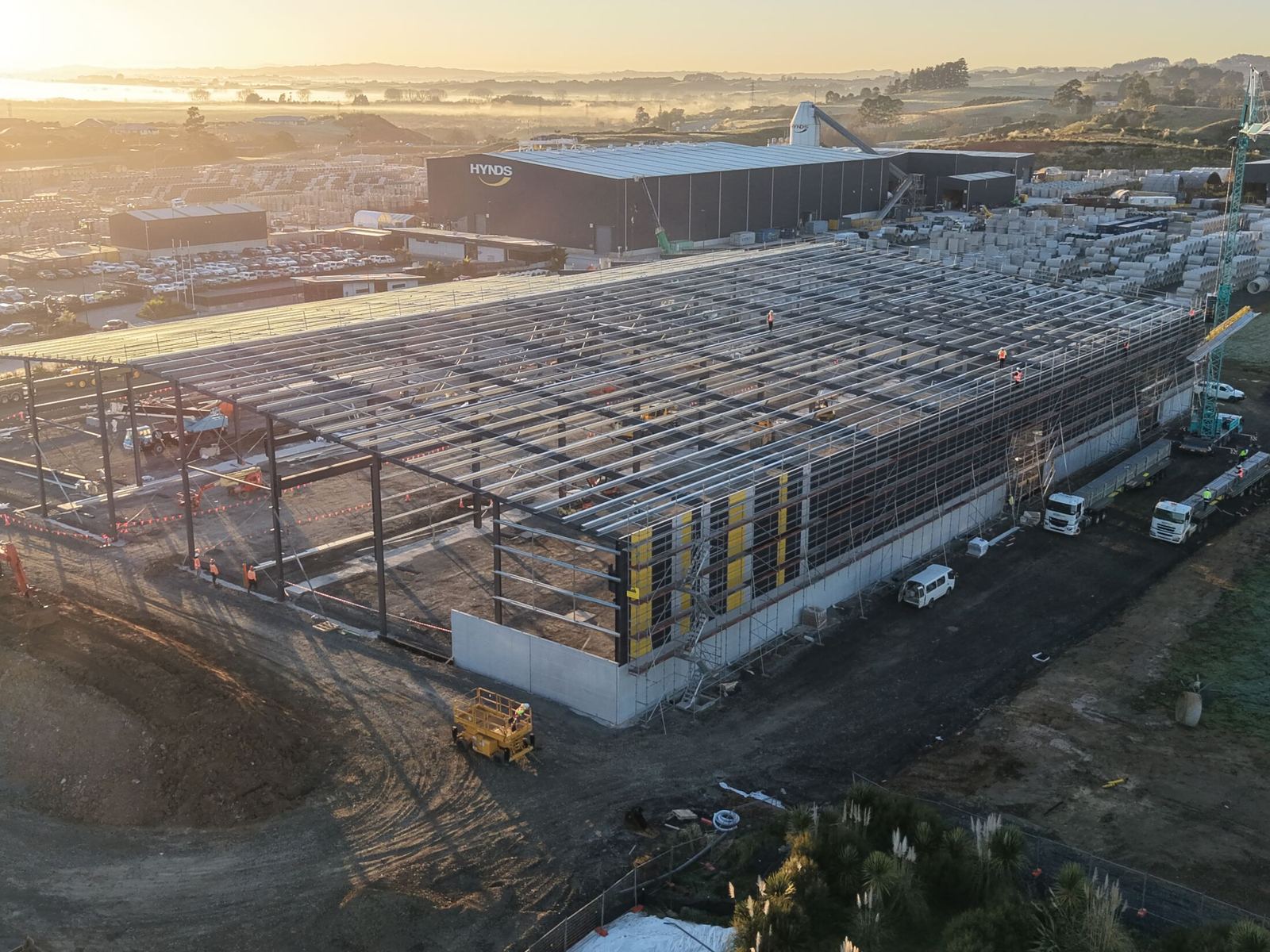 Dawn aerial of the Hynds Pōkeno manufacturing facility under construction, steel portal frames rising out of morning mist